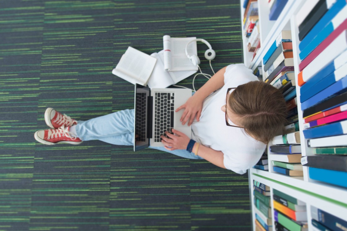 a person working on a laptop leaning against a bookshelf